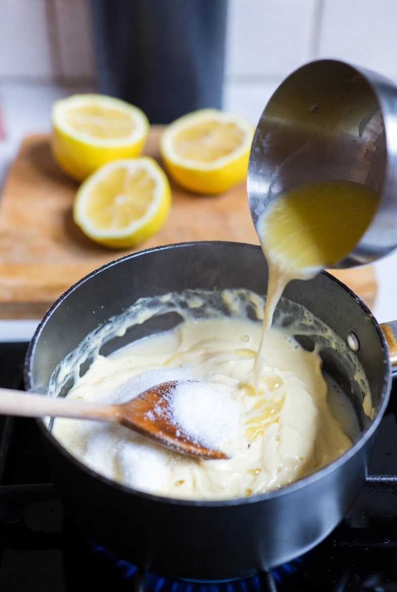 Cream mixture being stirred in a saucepan while making lemon posset with fresh lemon shells nearby